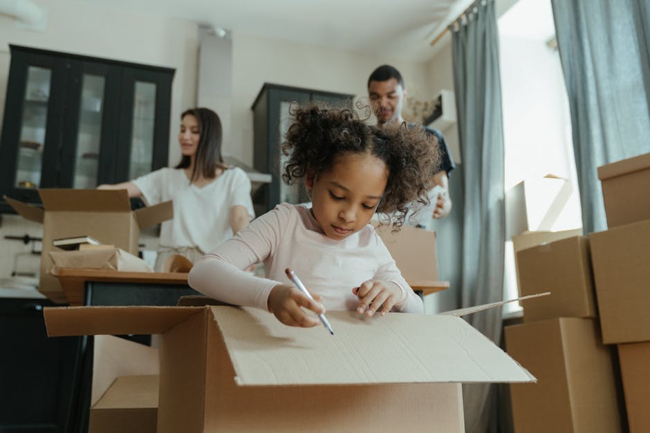 A family of five, consisting of two adult women, one man, and two children, is sitting on a wooden floor inside a home surrounded by packed cardboard moving boxes, some sealed with packing tape and ot