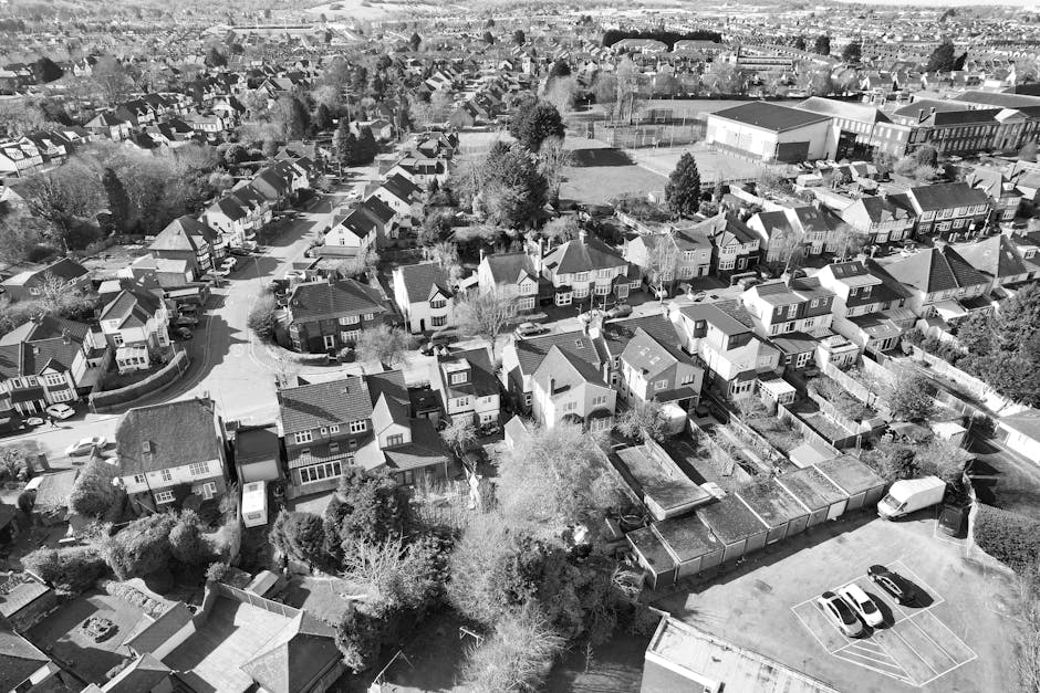 A black-and-white aerial photograph of a residential neighbourhood showing a variety of houses with pitched roofs, surrounded by trees and gardens. In the foreground, a move-in process is visible with a crew from Man with Van Barnehurst loading a wooden or cardboard box into a van, which is parked on the street near a driveway. The scene includes a small parking lot with a few parked cars, and the surrounding area features pavements, fences, and green spaces. The image depicts the logistics of home relocation, with focus on furniture transport, packing, and loading activities taking place during a house removal in Bexley. The lighting suggests it may be daytime with clear visibility of the property layouts and surroundings, illustrating typical moving operations in a residential area while aligning with local council permit rules for Barnehurst moving vans.