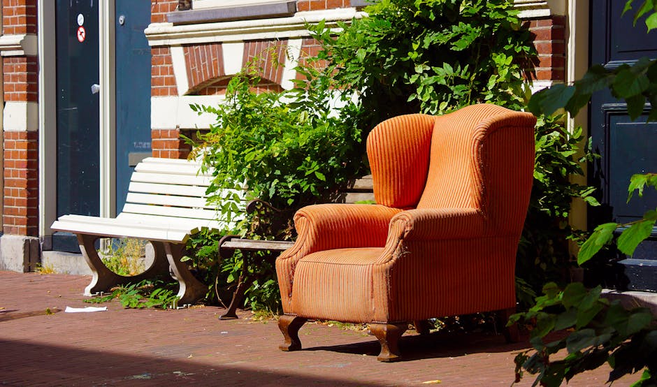 An outdoor scene showing an orange upholstered armchair with wooden legs positioned on a paved pavement in front of a brick building, partially surrounded by green foliage. Next to the armchair, there is a white wooden bench with curved legs, also placed on the pavement. The backdrop includes a dark blue door and a window with a white frame, indicating the entrance to a home. The natural lighting highlights the textures of the fabric on the chair, the brickwork, and the surrounding plants. This setting is related to home relocation and moving logistics, with items like furniture prepared for loading during a furniture transport or packing and moving process, as managed by Man with Van Barnehurst.