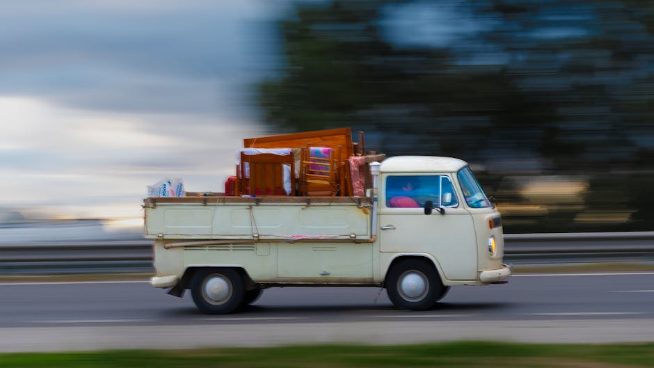 A small vintage white van with a boxy shape and rounded edges is shown in motion on a paved road, with a blurred background indicating speed. The van is loaded with various household items secured in the rear cargo area, including stacked wooden chairs, a cardboard box, plastic bags, and other miscellaneous furniture pieces covered with protective materials such as plastic wrap and fabric. The interior cabin is partially visible through the windshield, and the van is being used to facilitate a home relocation, with furniture and boxes prepared for transport. This scene illustrates furniture transport and loading procedures typical of professional removals services provided by Man with Van Barnehurst, as part of their bulk item removal offerings for house moves in the Barnehurst area.