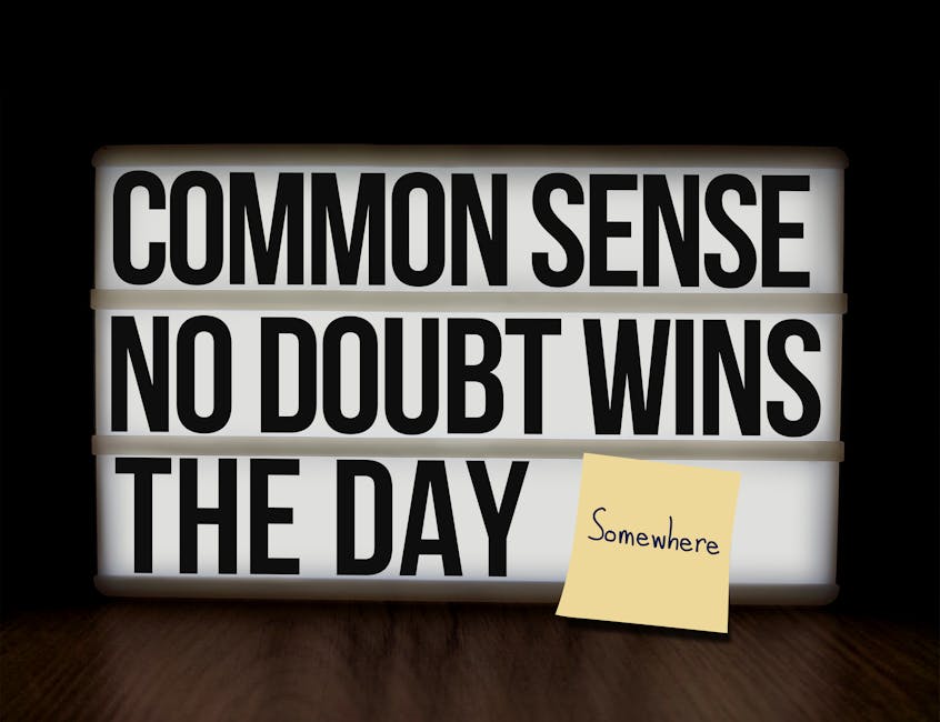 A backlit lightbox with a white frame displaying black uppercase letters that read 'COMMON SENSE NO DOUBT WINS THE DAY.' The lightbox is positioned on a dark wooden surface against a black background, illuminating the message. In front of the lightbox, a small yellow sticky note with handwritten text states 'Somewhere,' placed at a slight angle. The scene suggests a motivational or advisory message, with the setting resembling an indoor environment suitable for displaying quotes or notices related to moving or relocation planning, as seen in professional house removal services like those offered by Man with Van Barnehurst.