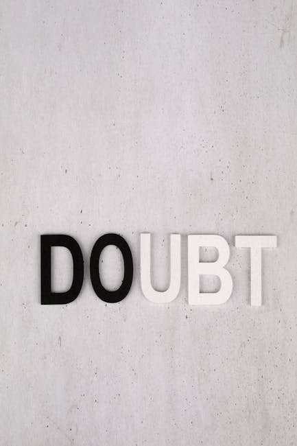 The image features the word 'Doubt' spelled out with individual three-dimensional letters mounted on a plain, light-colored concrete or plaster wall. The letters are arranged in a horizontal line, with the 'D' and 'B' in black, and the remaining letters in white, creating a visual contrast. The background wall has a smooth texture with subtle speckles or small imperfections. There are no other objects or furnishings visible in the scene. This visual could symbolise uncertainty or hesitation related to home relocation or moving decisions. In the context of house removals and packing services provided by Man with Van Barnehurst, it subtly underscores the importance of clear, professional relocation planning and transparent pricing during the moving process.