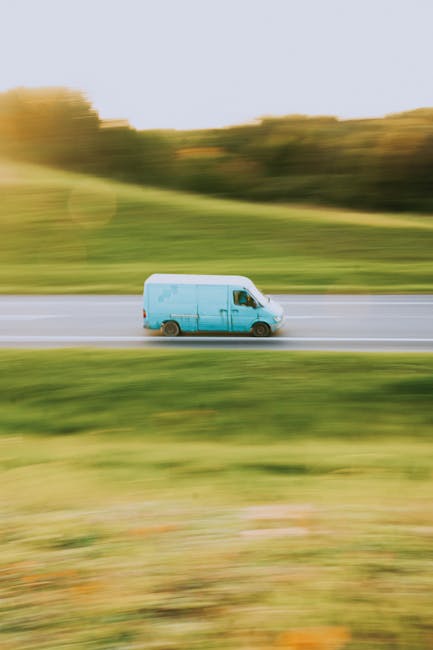 A moving blue cargo van is driving along a two-lane road in a rural area, with a grassy verge and yellow-orange trees in the background. The van appears to be in motion, with a slight blur indicating speed. Inside the van, visible through the driver's side window, are stacked cardboard boxes and wrapped furniture items, suggesting the vehicle is being used for home relocation or furniture transport. The scene is well-lit with natural daylight, and the surrounding environment includes a journey through open countryside, highlighting the logistical aspect of house removals. This image relates to moving services offered by Man with Van Barnehurst, illustrating the transportation of household items during a furniture moving process on a typical route for home relocations like Crook Log to Barnehurst.