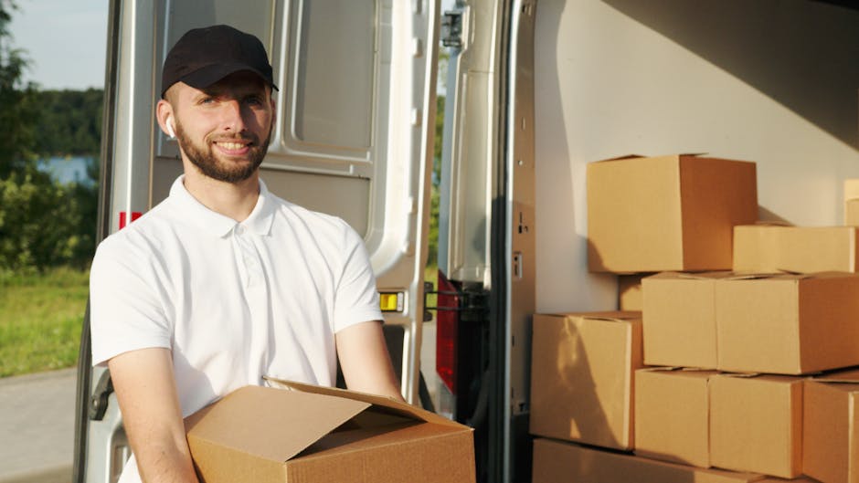 A man wearing a white shirt and sunglasses is seen loading a cardboard box into the back of a moving van during a home relocation. Inside the van, several other cardboard boxes of varying sizes are vi