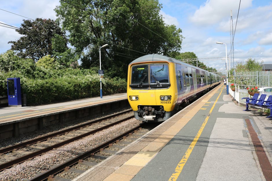 A modern yellow and grey train is arriving at an outdoor station platform during daytime, with its front facing the viewer. The platform features a yellow safety line near the edge, blue seating benches on the right, and a white metal fence decorated with flower pots. In the background, there are lush green trees and a partly cloudy sky. Overhead electric lines run parallel to the train tracks, which are embedded in gravel. On the left side of the image, there is a blue public telephone booth and a tactile paving strip for visually impaired passengers. The station environment appears clean and well-maintained, indicating an urban or suburban setting suitable for house and furniture removals, with the train station serving as a key part of local transport logistics facilitated by companies like Man with Van Barnehurst.