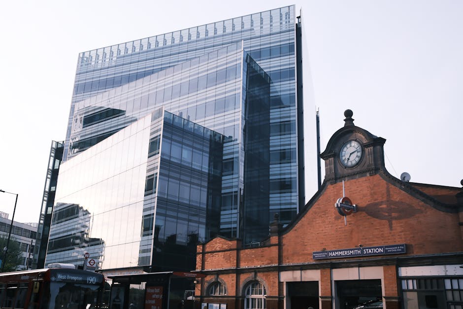 The image shows a modern, multi-storey glass office building with reflective windows, adjacent to an older brick railway station structure labeled 'Hammersmith Station' with a traditional clock mounted on its facade. In the foreground, a white van with the signage of Man with Van Barnehurst is parked on the street, partially visible near the station entrance. The van appears to be involved in the loading process, as part of a house removal or home relocation service. The scene captures the urban environment during daytime with clear lighting, highlighting the contrast between contemporary commercial architecture and historical railway elements, supporting the context of local moving and furniture transport services near a busy city station.