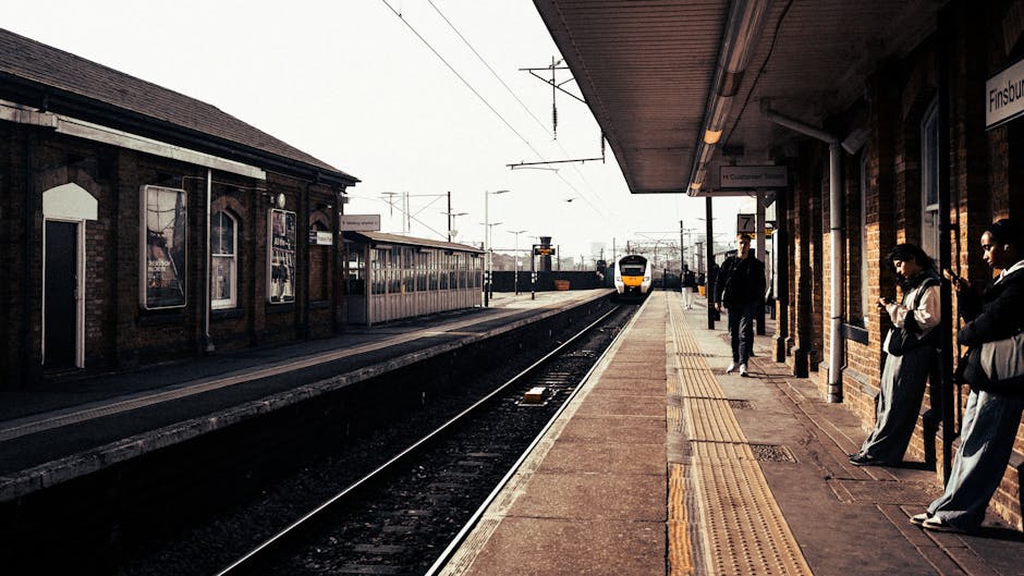 A view of a train station platform during daytime with several people waiting for a train. On the platform's right side, three individuals are standing near the brick wall, some reading or looking at their phones, while one person appears to be preparing to board. The platform is equipped with tactile paving along the edge for safety, and overhead lighting fixtures are visible under the station's roof. A yellow and white passenger train is approaching in the distance on the railway tracks, indicating active train services. Across the tracks, there is a small station building with windows and advertising posters on the brick exterior. The overall scene captures a typical railway station environment, relevant to home relocation or moving services such as those offered by Man with Van Barnehurst, emphasizing the logistics of furniture transport and moving logistics associated with train-based travel and station access.