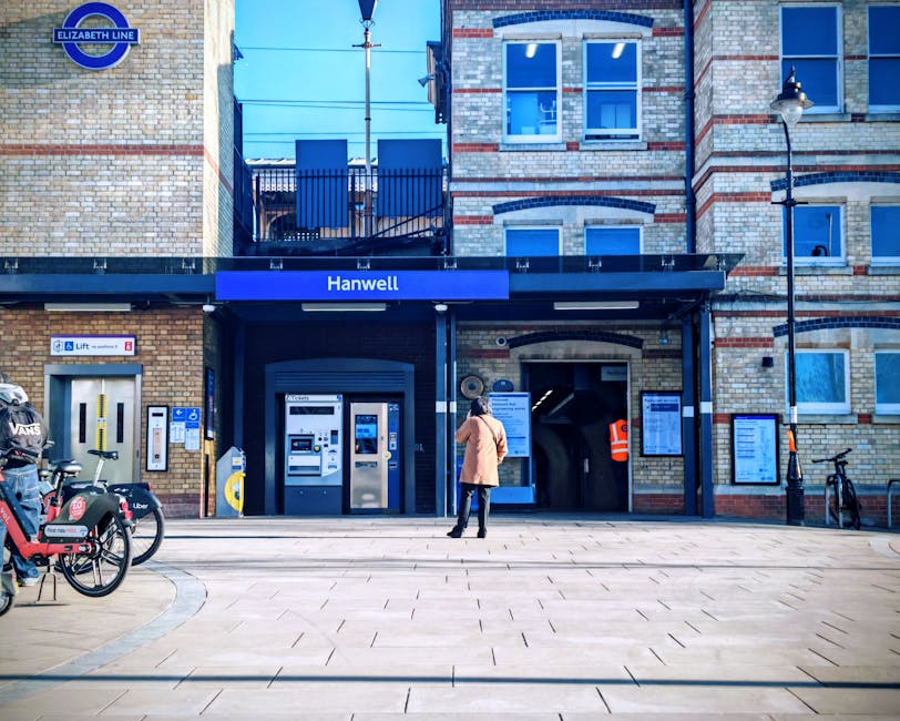 A woman stands outside Hanwell station, part of the Elizabeth Line, in front of the station entrance which features a dark canopy with the station name displayed on a blue sign. To her left, there are two rental bicycles secured to a bike stand, and a pair of vending machines, one for tickets and the other for snacks or drinks. The station building has a brick façade with windows above and a door to the right of the entrance. A lamppost and a bicycle are also visible on the right side. The scene is illuminated by daylight, and the pavement in front of the station is clean and spacious. This setting provides context for local transportation options relevant to house removals and furniture transport in connection with moving services offered by Man with Van Barnehurst, supporting home relocation logistics.