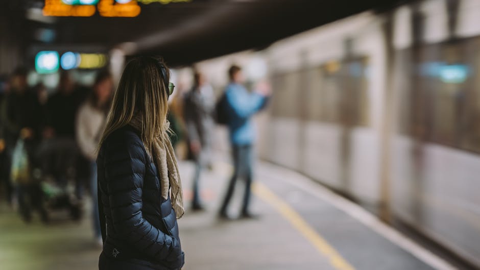A view of a train station platform during daytime with several people waiting for a train. On the platform's right side, three individuals are standing near the brick wall, some reading or looking at their phones, while one person appears to be preparing to board. The platform is equipped with tactile paving along the edge for safety, and overhead lighting fixtures are visible under the station's roof. A yellow and white passenger train is approaching in the distance on the railway tracks, indicating active train services. Across the tracks, there is a small station building with windows and advertising posters on the brick exterior. The overall scene captures a typical railway station environment, relevant to home relocation or moving services such as those offered by Man with Van Barnehurst, emphasizing the logistics of furniture transport and moving logistics associated with train-based travel and station access.
