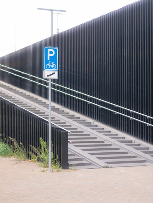 Image showing a set of outdoor ramp access with metal steps and handrails leading up to a building, situated beside a tall black slatted fence. A blue parking sign with a bicycle symbol and a directional arrow is mounted on a pole next to the ramp, indicating designated parking for cyclists. The ramp is composed of concrete or metal with ridged surfaces for traction, and the setting appears to be part of an urban or commercial area. In the foreground, there is a paved pathway with some greenery along the edge. The scene is well-lit with natural daylight, emphasizing the functional design of the staircase and access pathway, relevant to logistics, house removals, and furniture transport considerations addressed by Man with Van Barnehurst.