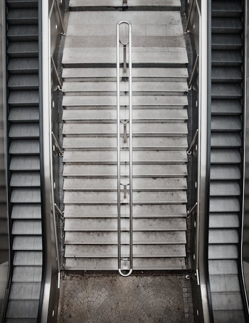 A top-down view of an indoor staircase with metal handrails and concrete steps, situated between two escalators with black rubber steps. The staircase begins at the ground floor, which has a tiled circular pattern, and leads upward. The concrete steps are evenly spaced and have a smooth surface, with a metal handrail running down the center of the staircase for safety. The area is illuminated by natural or artificial light, highlighting the utilitarian design. This staircase is part of a building's interior, likely a commercial or residential property, and is adjacent to escalators that facilitate movement between different levels. This scene is related to house removal and moving logistics, illustrating potential access issues or practical fixes for staircases in property relocation scenarios, as addressed by Man with Van Barnehurst's removals services.