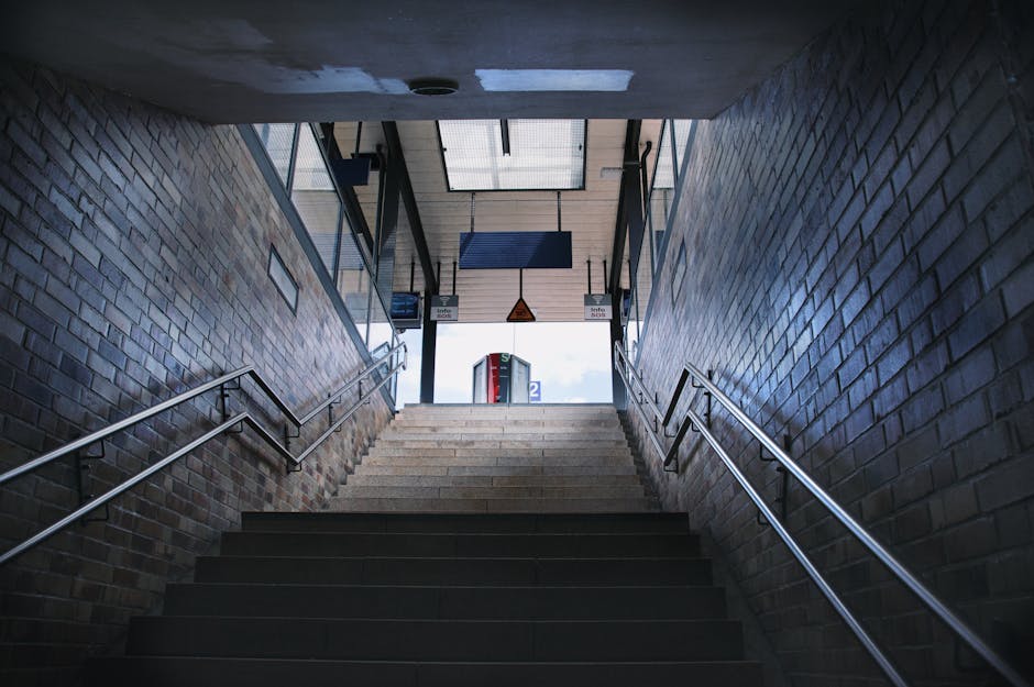 Image showing a set of outdoor ramp access with metal steps and handrails leading up to a building, situated beside a tall black slatted fence. A blue parking sign with a bicycle symbol and a directional arrow is mounted on a pole next to the ramp, indicating designated parking for cyclists. The ramp is composed of concrete or metal with ridged surfaces for traction, and the setting appears to be part of an urban or commercial area. In the foreground, there is a paved pathway with some greenery along the edge. The scene is well-lit with natural daylight, emphasizing the functional design of the staircase and access pathway, relevant to logistics, house removals, and furniture transport considerations addressed by Man with Van Barnehurst.