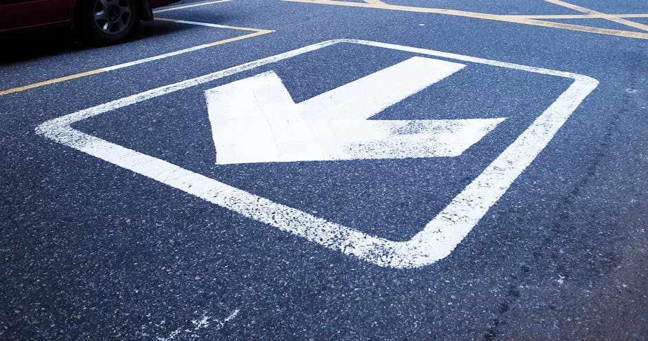A close-up view of a designated loading bay painted on an asphalt pavement, featuring a white downward-pointing arrow enclosed within a square outline. The surface is textured with small gravel particles and the white paint appears slightly worn, indicating frequent use. In the background, part of a black vehicle is visible on the curb, suggesting this area is used for vehicle loading and unloading during home relocations or furniture transport. The setting reflects an organized space for moving services, such as those offered by Man with Van Barnehurst, to facilitate packing and loading processes in tight street conditions like Station Road, DA7, where careful navigation is essential for efficient removals.
