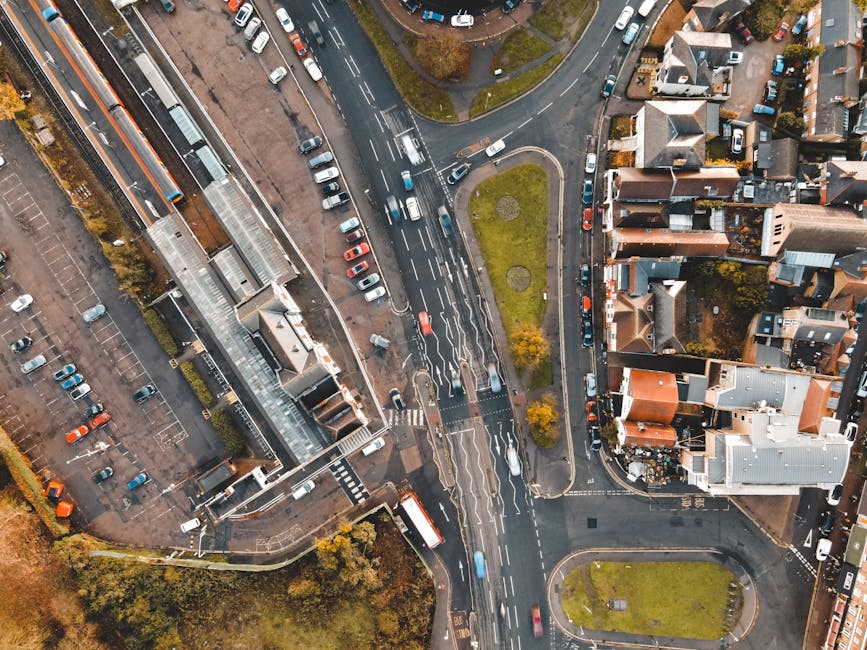 An aerial view of a residential street showing the loading process for a home relocation, with a Man with Van Barnehurst vehicle parked on the pavement. Several cardboard boxes and wrapped furniture are visible on the roadside, ready to be loaded into the van. The image depicts a small group of movers, equipped with blankets and straps, carefully moving furniture and boxes from the property to the vehicle. The surrounding area includes tightly packed houses with front gardens, parked cars along the curb, and a small central traffic island with grass and a few trees. Sunlight illuminates the scene, highlighting the logistics involved in household packing and furniture transport during a house removal, carried out within the narrow streets of the neighbourhood.