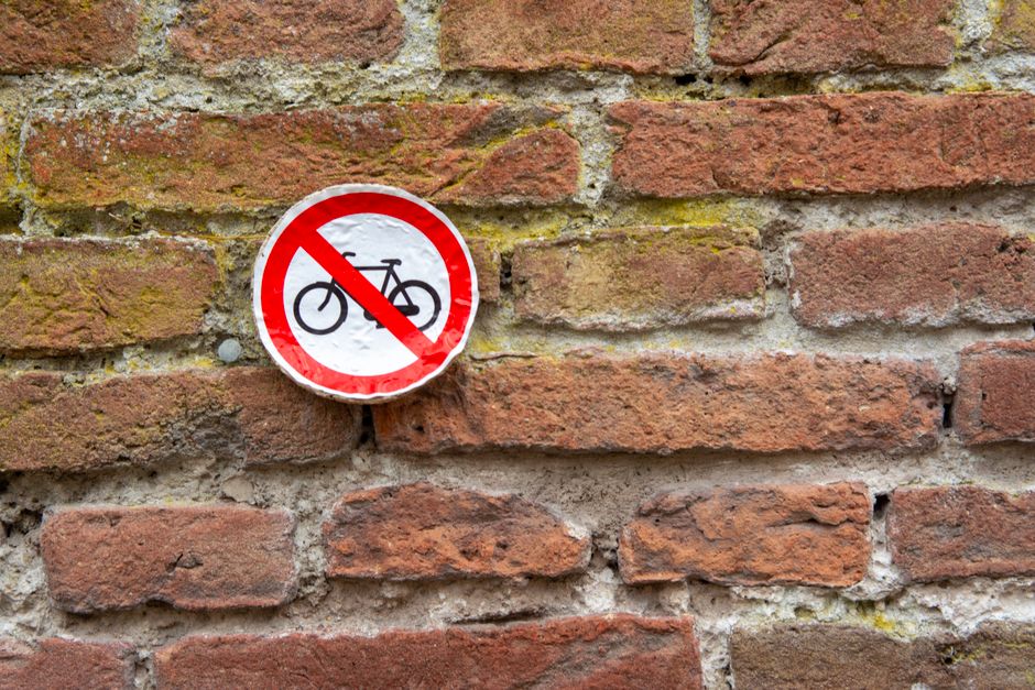 A circular traffic sign with a red border and a white background, displaying a black bicycle symbol crossed out with a red diagonal line, mounted on a weathered brick wall. The bricks are reddish-brown with some moss and mortar visible between them. The sign is positioned approximately at the middle height of the wall, indicating that bicycles are not permitted in the area. The wall surface shows signs of age, with uneven textures and slight discoloration. This image relates to household or local authority regulations concerning street use and parking, which may be relevant for home relocation or moving logistics when planning furniture transport or clearance in the vicinity. Man with Van Barnehurst offers removals services that include navigating local rules, such as restrictions on bicycle access, during house moves or furniture transport, as seen in this urban environment.
